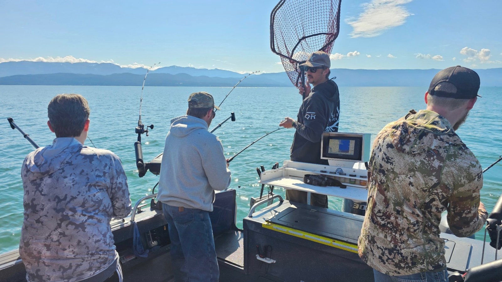 Four people fishing on a boat, one holding a large net, calm lake and mountains in the background.