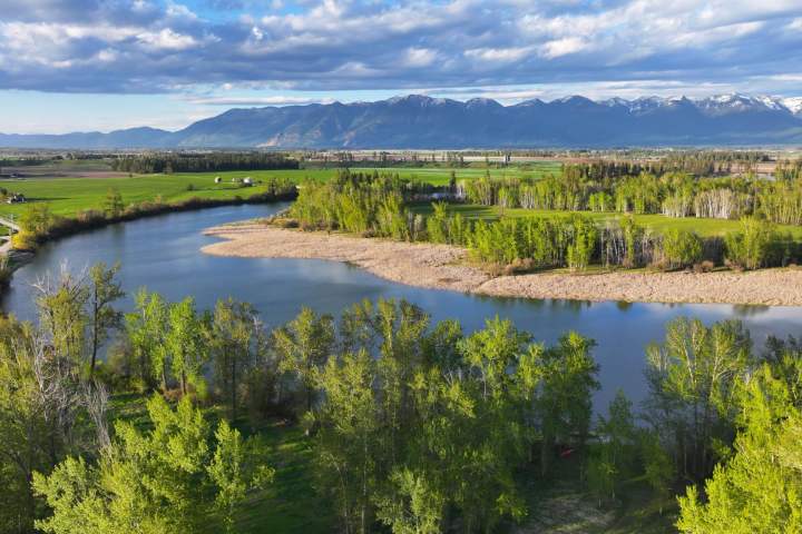 Scenic landscape with a river, green fields, and mountains under a cloudy sky.