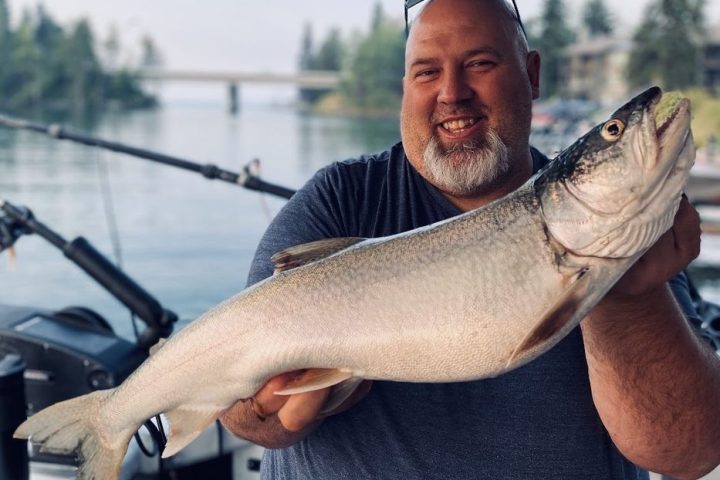 Smiling man on a boat holding a large fish with rods in background.