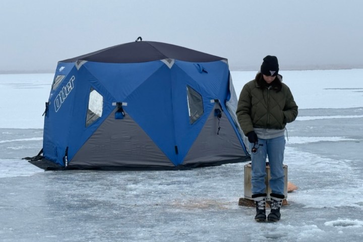 Person standing on ice near a blue tent on a frozen lake.