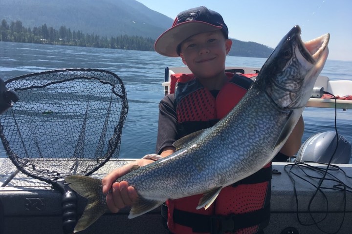 Child on boat holding a large fish, with mountains and lake in background.
