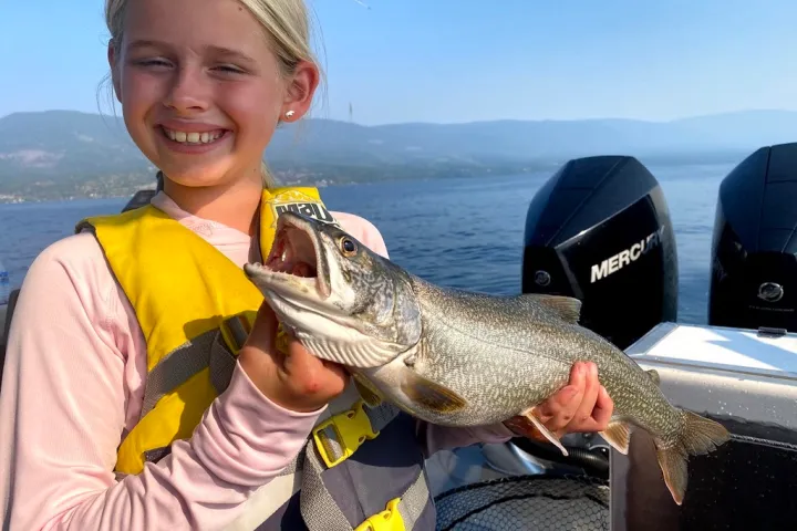 Child smiling on a boat holding a large fish, with mountains and water in the background.