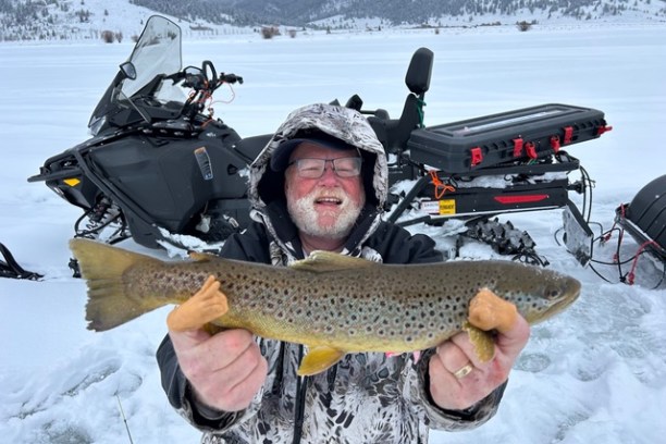 Man holding a brown trout on a snowy landscape with snowmobile.