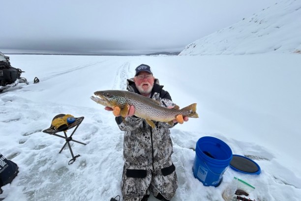 Person kneeling on ice holding a large fish, surrounded by fishing gear and snow.