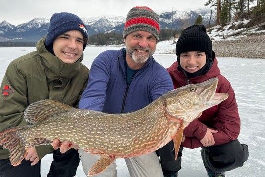 Three people on ice, holding a large fish, mountains in the background.