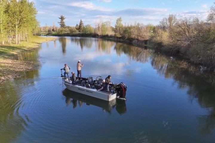 Two people fishing on a boat in a calm, tree-lined river.