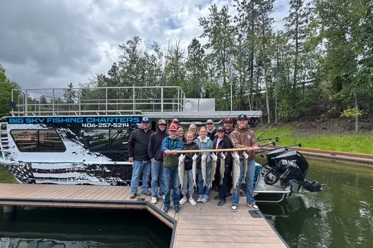 Group on dock holding caught fish, with a boat and trees in background.