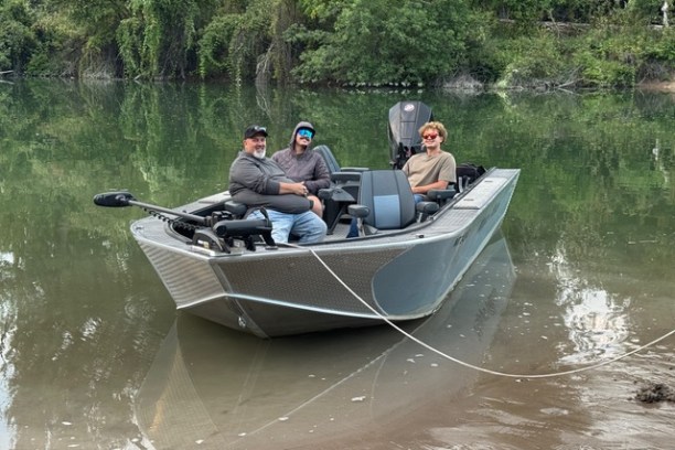Three people sitting in a boat on a calm river near the shore with trees in the background.