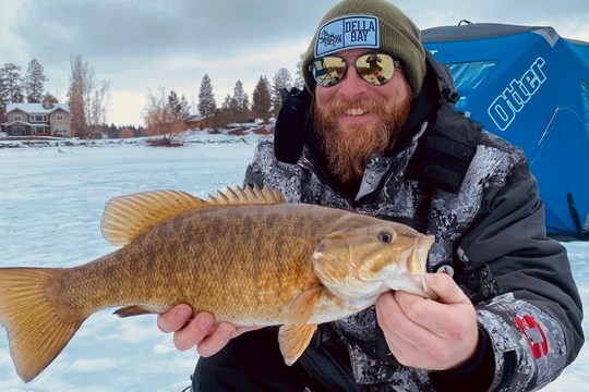Person holding a fish while ice fishing, wearing winter clothing and sunglasses, icy landscape in background.