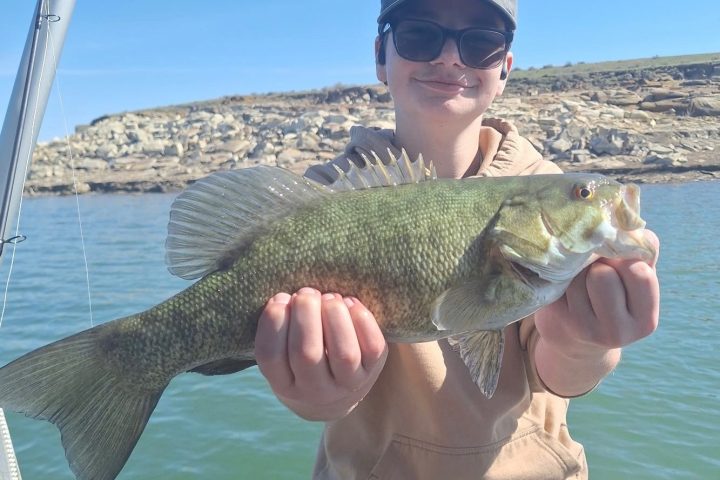 Person holding a large fish on a boat, with rocky shoreline in the background.