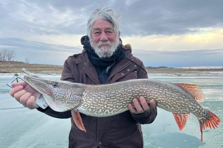 Man holding a large fish on a frozen lake with cloudy sky.