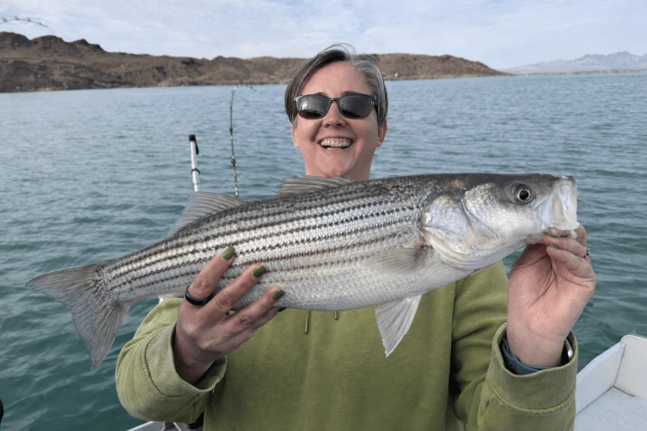 Person smiling and holding a large fish on a boat with a lake and hills in the background.