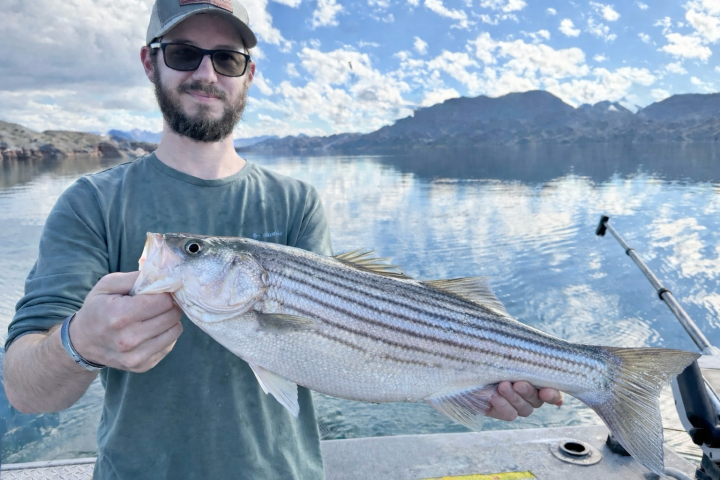 Man holding a large fish on a boat with a lake and mountains in the background.