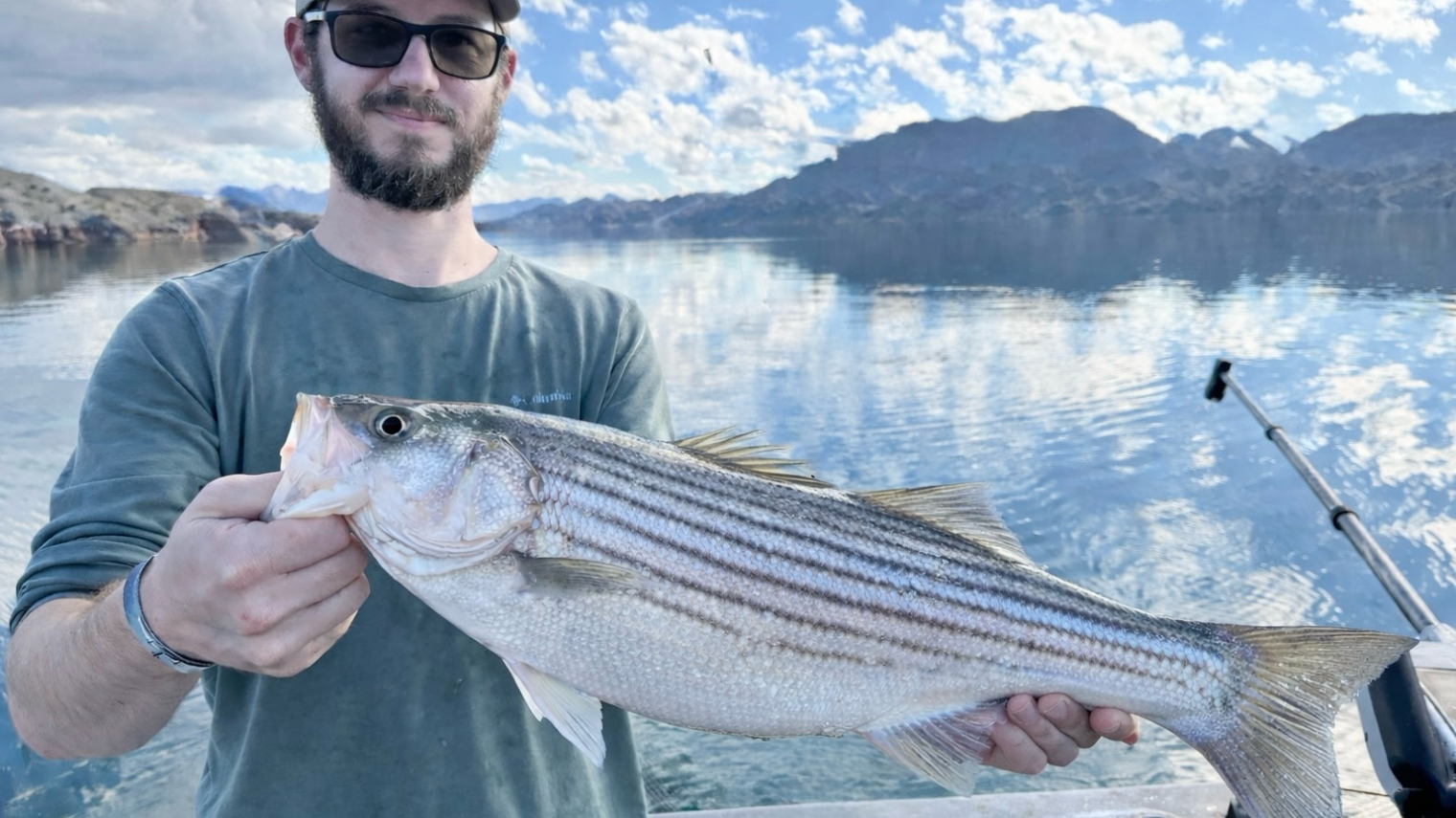 Man holding a large fish on a boat with a lake and mountains in the background.